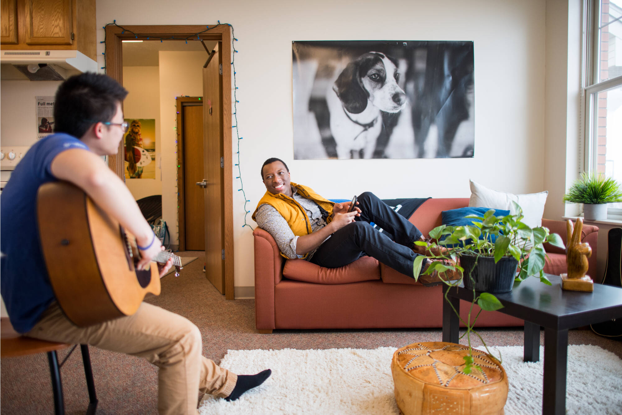 Two people hang out in a VanSteeland living room. One person plays guitar while the other lounges on an orange couch, smiling.